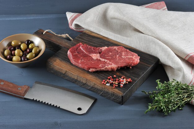 Piece of raw beef on a wooden Board, a knife for meat, olives and thyme