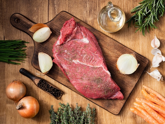 A piece of raw red meat lies on a vintage cutting board with green herbs. Healthy protein. Pepper beans. White onions.