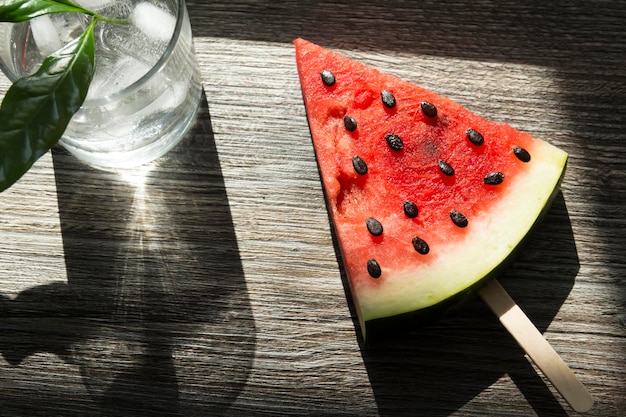 A piece of triangular watermelon on an ice cream stick and a glass of cold drink with ice