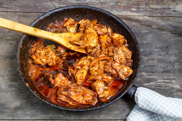 Pieces of baked chicken in sauce in a frying pan with a wooden spatula on a dark table