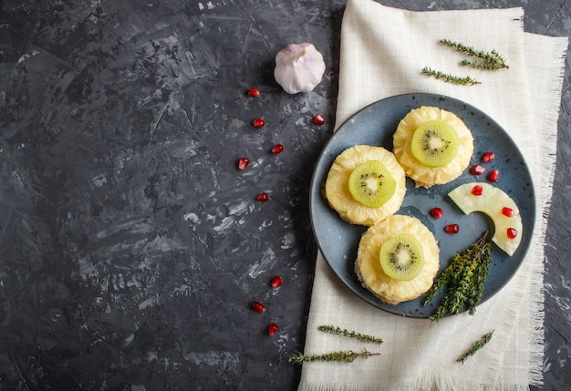 Pezzi di carne di maiale al forno con ananas, formaggio e kiwi su sfondo nero.