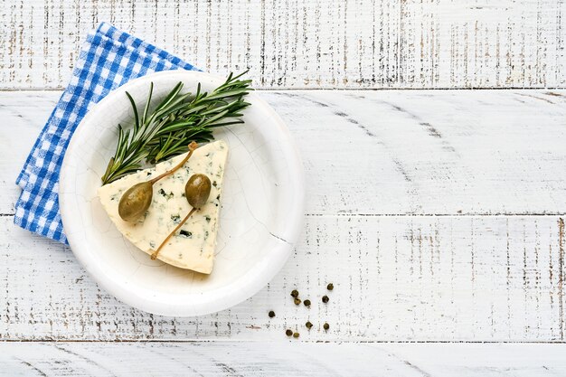 Pieces of blue cheese or brie cheese in a bowl with rosemary, capers and pepper. 