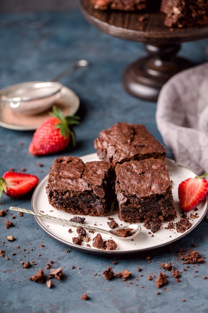 Pieces of chocolate nut brownie, on white plate, spoon, with slices of strawberries, crumbs, grey textile. Dark blue background. Vertical