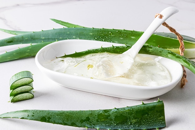 Pieces of a cut layer of aloe in a white oval bowl among the\
leaves of the plant on a marble background the concept of preparing\
the gel at home