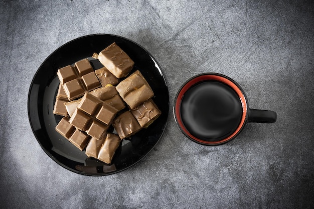 Pieces of different chocolate on black dish and cup of coffee with steam on gray table view above