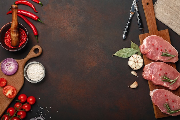 Фото Кусочки сырой свиной стейки на разделочной доске с помидорами черри, розмарином, чесноком, красным перцем, лавровым листом, луком