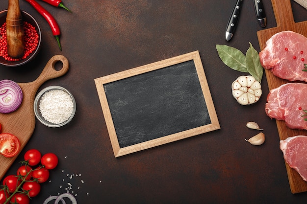 Pieces of raw pork steak on cutting board with cherry tomatoes, rosemary, garlic, red pepper, bay leaf, onion