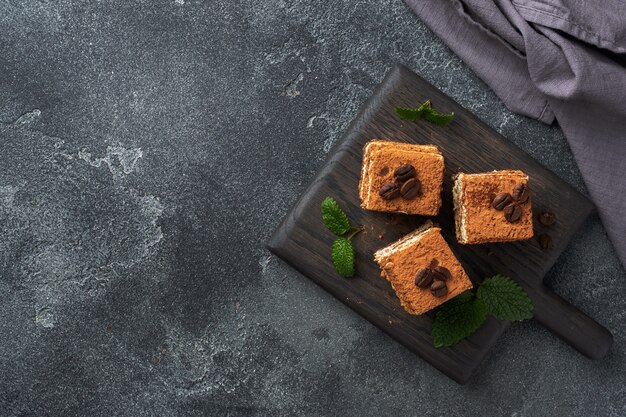 Pieces of tiramisu cake with delicate cream, coffee beans and mint leaves. Dark concrete background with copy space. Top view.