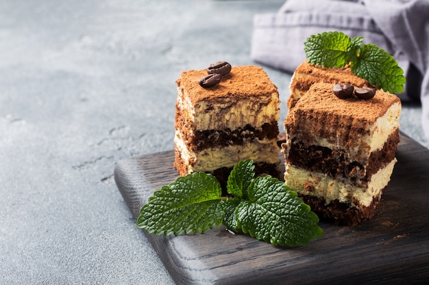 Pieces of tiramisu cake with delicate cream, coffee beans and mint leaves. Dark concrete table with copy space.