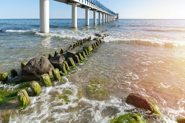 Pier aan kust. Surf golven met zeeschuim en zeewier op toeristenstrand. Mooie kust in zonlicht. Dag zeegezicht.