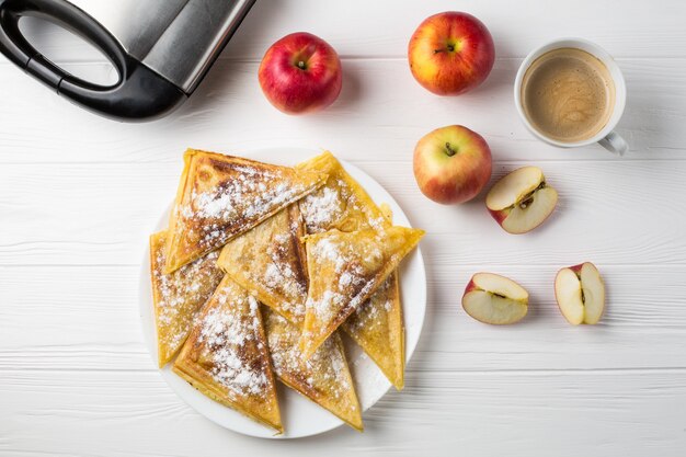 Pies lie on the table next to apples, a toaster and a cup of coffee