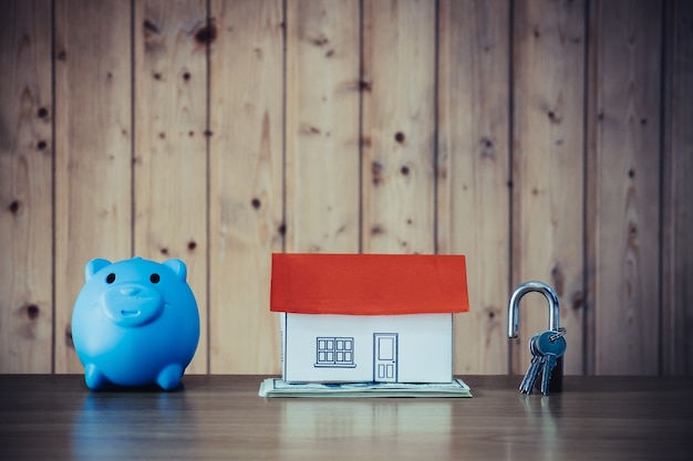 Pig money box and paper house with key on wooden wall background