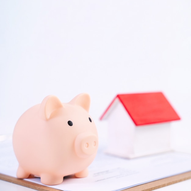 Piggy bank, beautiful wooden house model over a signed contract on white background, concept of saving money to buy a home insurance, close up, copy space.