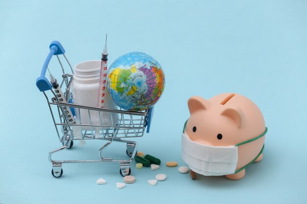 Piggy bank in a medical mask and shopping trolley with pills bottle, syringe on blue background. Covid-19 pandemic