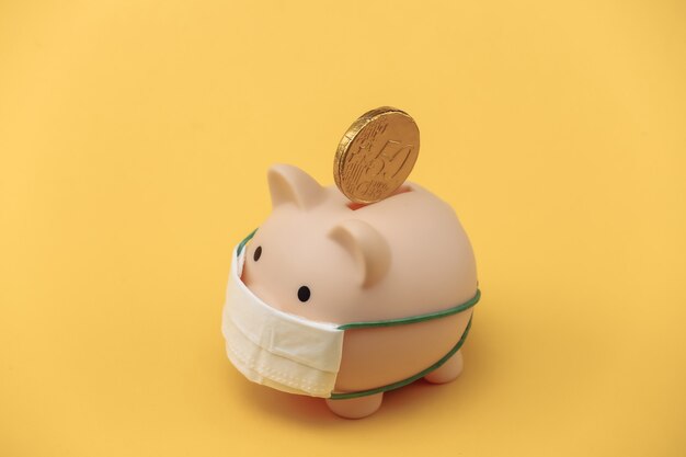 Piggy bank in medical mask with coin on yellow background. Economic disease. Financial crisis. Covid-19 pandemic