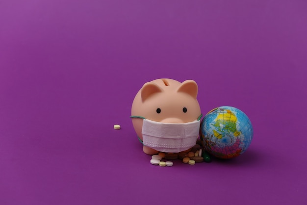 Piggy bank in medical mask with globe, pills on purple background. Covid-19 pandemic