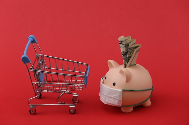 Photo piggy bank in a medical mask with a shopping trolley on red background. shopping during covid-19, economic crisis, economy