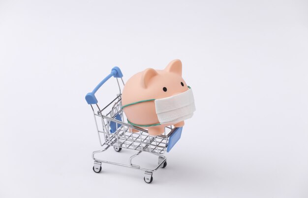 Piggy bank in a medical mask with a shopping trolley on white background. Shopping during covid-19, economic crisis, economy