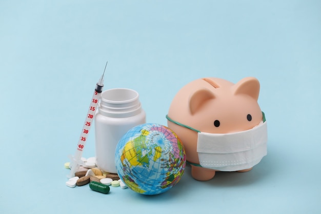 Piggy bank in medical mask with syringe, pills bottle on blue background. Vaccination. Covid-19 treatment. Global pandemic