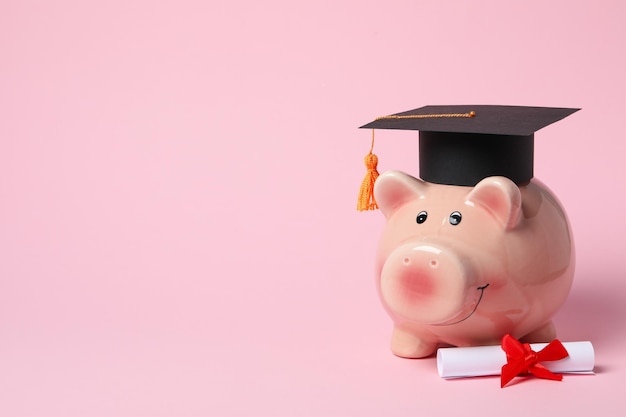 Piggy bank with graduation hat and degree scroll on pink background