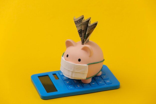 Piggy bank with medical mask and calculator on yellow background. Economic disease. Financial crisis. Covid-19 pandemic