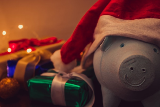 Piggy bank with Santa Claus hat on blurred christmas background with christmas ornaments, gifts and light. Space for text.