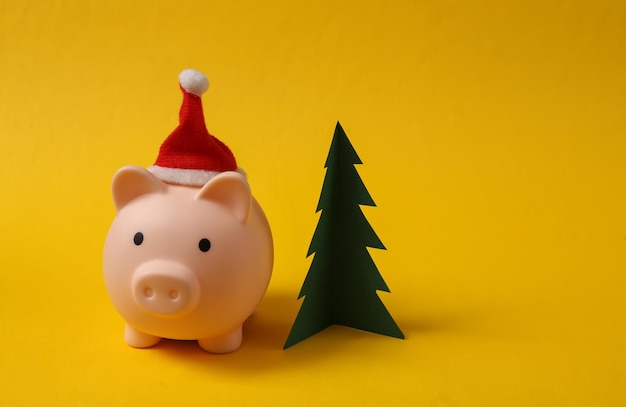 Piggy bank with santa hat and Christmas tree on yellow background Christmas concept