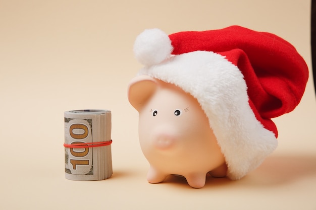 Piggy money bank with Christmas hat bunch of dollars banknotes cash money isolated on beige background. Money accumulation investment, banking services, wealth concept. Copy space advertising mock up.