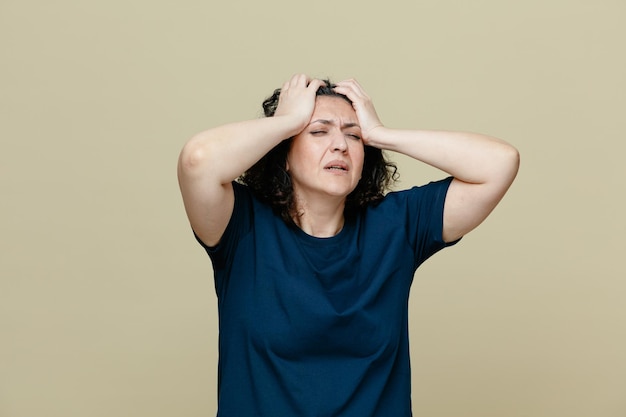 Pijnlijke vrouw van middelbare leeftijd met een t-shirt die de handen op het hoofd houdt met gesloten ogen met hoofdpijn geïsoleerd op een olijfgroene achtergrond