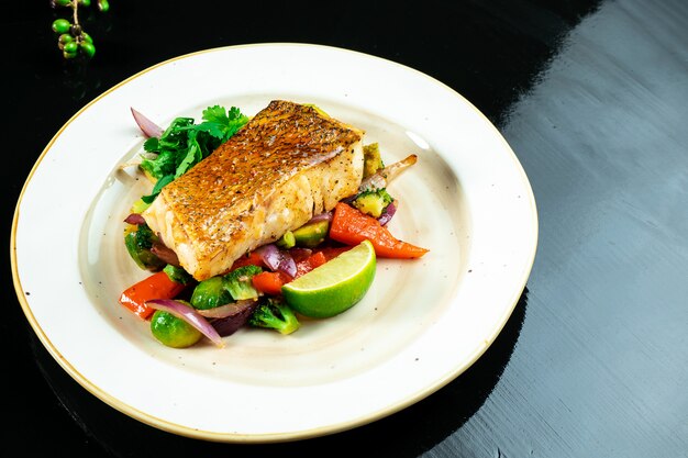 Pike perch fillet in spicy sweet sauce on a pillow of boiled vegetables in yellow plate on dark background.