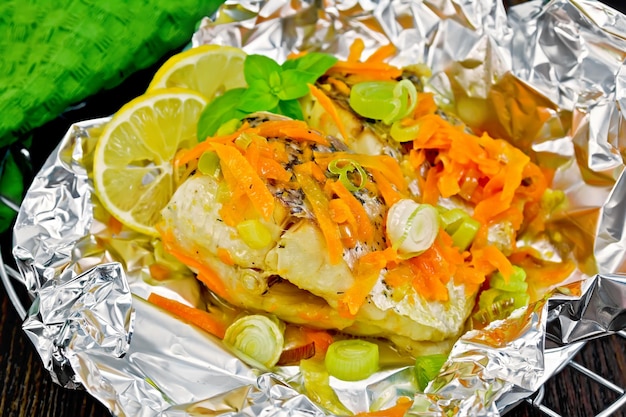 Photo pike with carrots, leek, basil and slices of lemon in foil on the lattice, towel on a background of dark wood planks