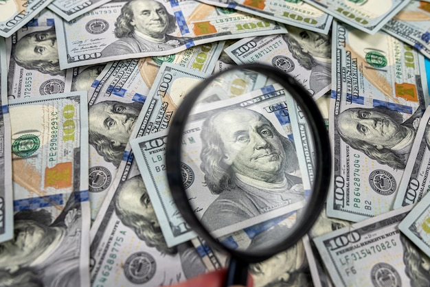 Photo pile of hundred dollar bills with a portrait of president benjamin franklin under a magnifying glass