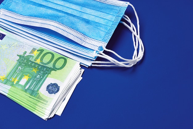 Pile of medical face masks and Euro banknotes on blue background