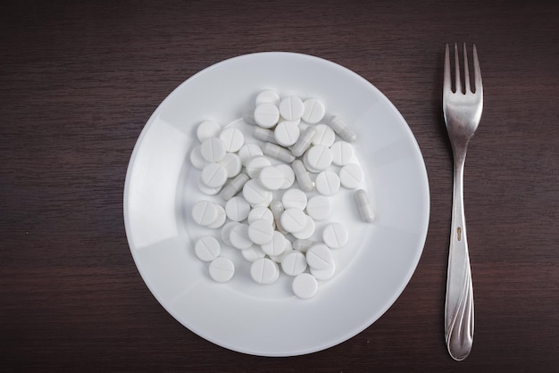A pile of medical pills with a fork on a plate on a wooden\
table. pill overdose concept. the use of dietary supplements. top\
view. flatly.