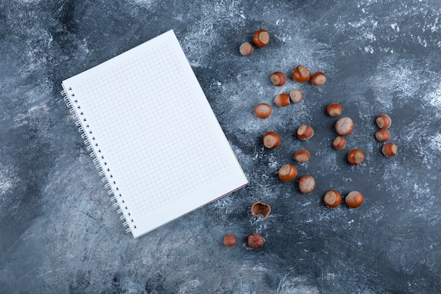 Pile of organic shelled hazelnuts with empty notebook on marble.