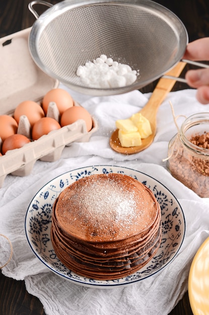 A pile of pancakes with a wooden spoon of butter, eggs, on a rustic wooden table.