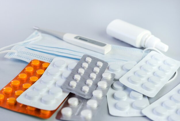 Pile of pills in blister packs, medical thermometer and mask on gray background. Disease prevention and treatment concept. Top view. Close up.