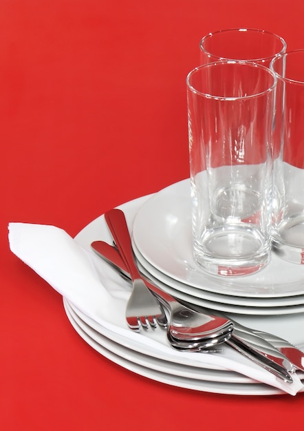 Photo pile of white plates, glasses with forks and spoons on silk napkin. red background