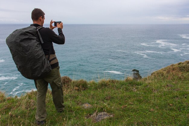 Pilgrim taking picture on ocean coast Tourist with backpack on hill Man on aerial seascape