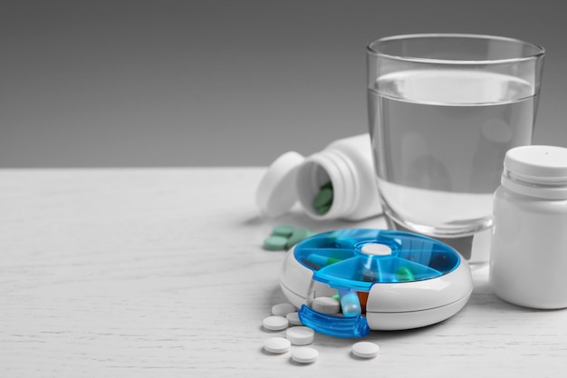 Pill box with medicaments and glass of water on white wooden table Space for text