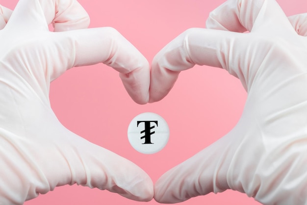 A pill with a tugrik currency sign inside a heart made from hands in medical gloves on a pink background Rise in the price of medicines in Mongolia Expensive treatment