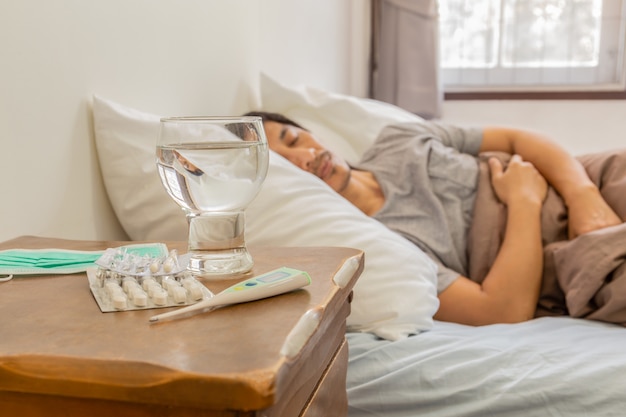 錠剤、コップ一杯の水、温度計、ベッドで寝ている男の横にある錠剤。