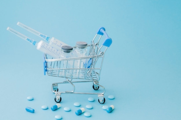 薬と青の背景にショッピングトロリーで医療注射。医療費、ドラッグストア、健康保険、製薬会社のビジネスコンセプトの創造的なアイデア。コピースペース