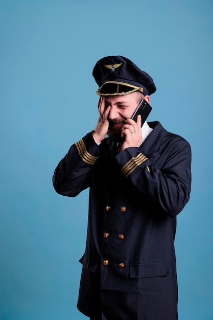 Piloot van middelbare leeftijd die op smartphone praat met gezichtspalmuitdrukking, mobiele telefoon vasthoudt, een gesprek voert. Luchtvaartkapitein in uniform staand, telefoon beantwoordend