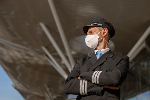 Pilot in protective mask in uniform standing outdoors near airport building