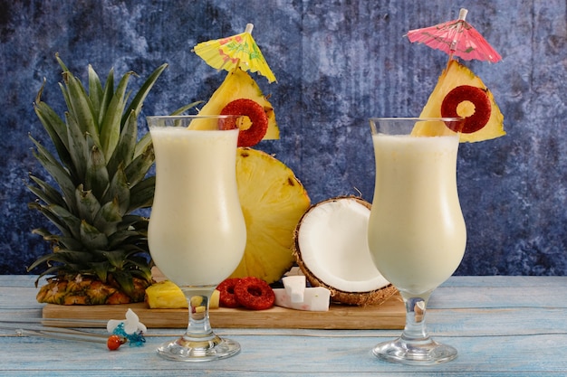 Piñas coladas with fresh fruits on wooden cutting board on white board table and blue background