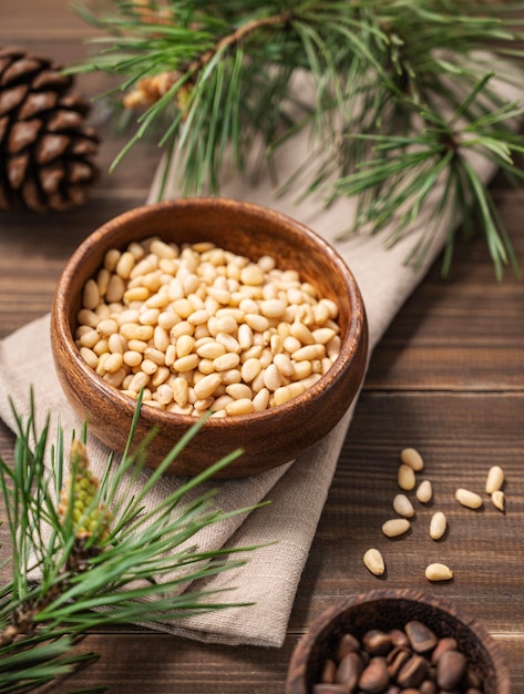 Pine nuts in a bowl on napkin and a handful of unpeeled nuts on a brown wooden background with a branch of pine needles Healthy diet snack
