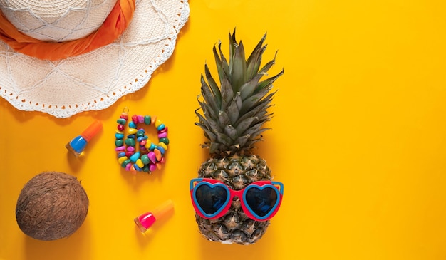 Foto ananas in occhiali da sole a forma di cuore, cappello di paglia e altri accessori estivi su sfondo giallo