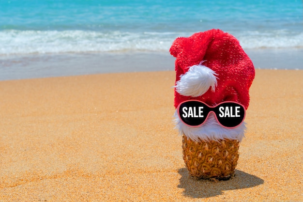 熱帯のビーチでテキスト販売とメガネのサンタクロースの帽子のパイナップル。ブラックフライデー