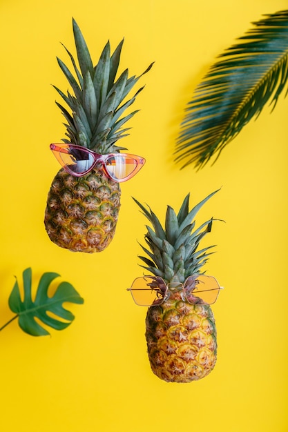 Pineapples creative faces in color sunglasses with palm leaves on yellow color summer background. Tropical summer fruits levitating pineapples.